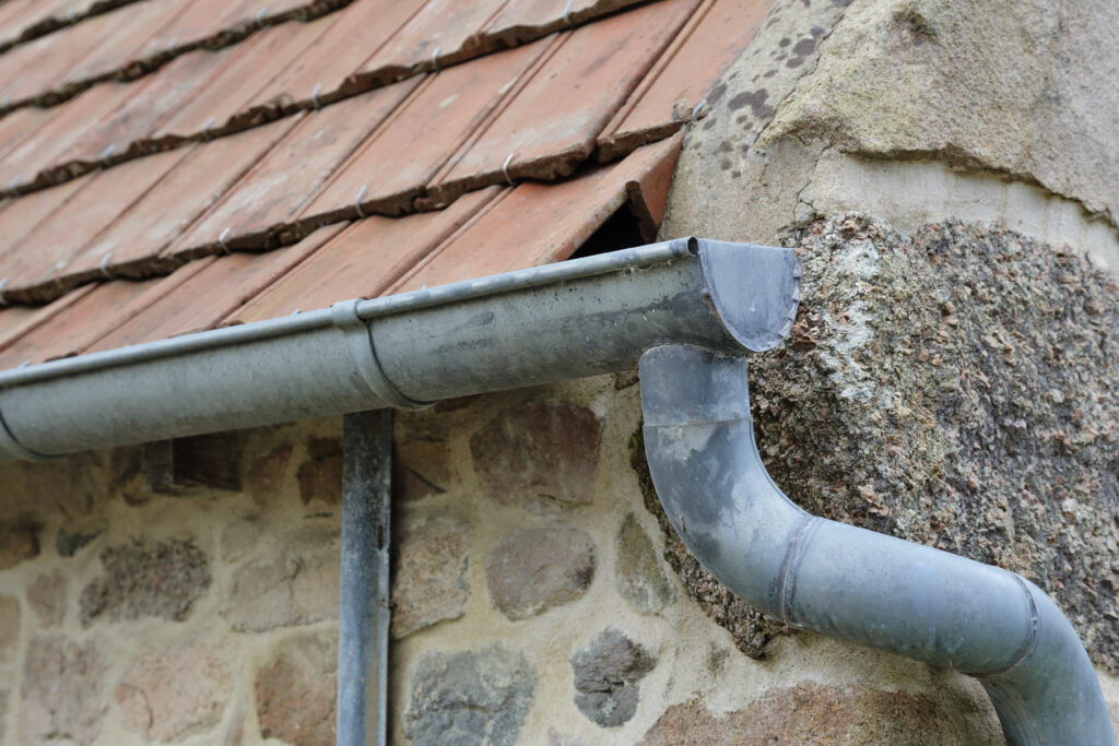 Gouttière et toit de tuiles : Détail architectural rustique Gouttière métallique grise sur un mur en pierre rustique, sous un toit de tuiles en terre cuite. Des crochets maintiennent les tuiles.